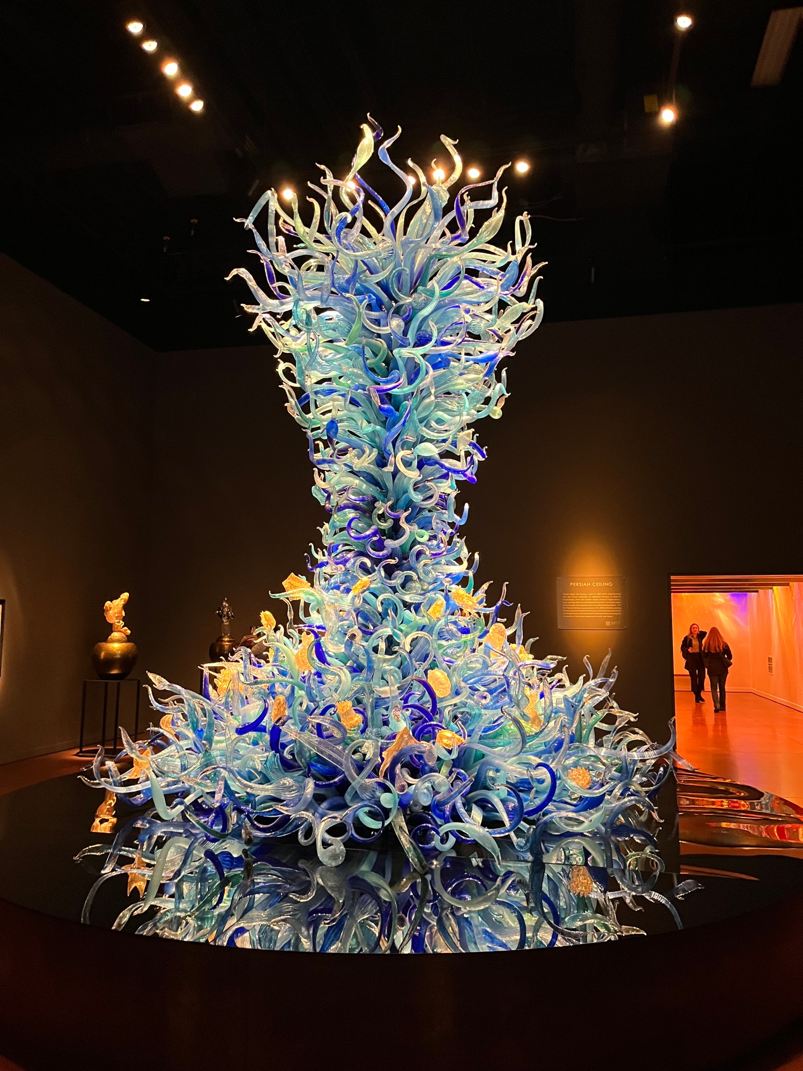 A floor to ceiling glass sculpture by Dale Chihuly at the Seattle glass museum. Each piece of glass is a large tendril coming out of a central tornado-like structure. The pieces of glass are various hues of blue. There are gold glass sculptures at the bottom of starfish and nautilus.