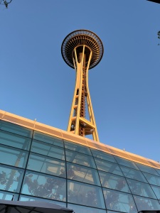 The picture of the Space Needle in Seattle. The sky is clear blue, there's a greenhouse building in the foreground. The photo is taken from the ground looking up.