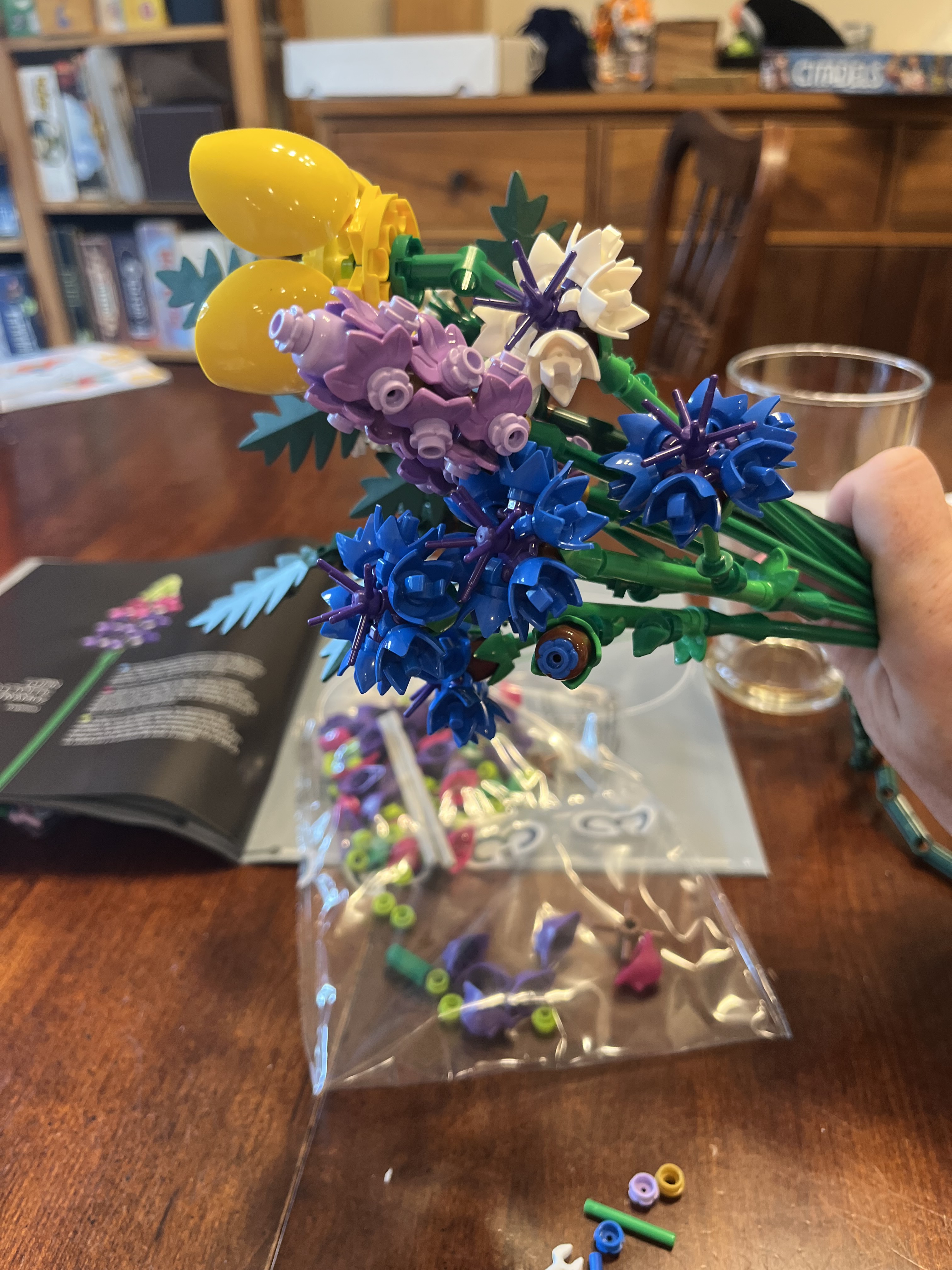 In the foreground is a collection of colorful Legos constructed into wildflowers. A hand on the right is holding the bunch of flowers. In the background is a dining table and the Lego instruction book